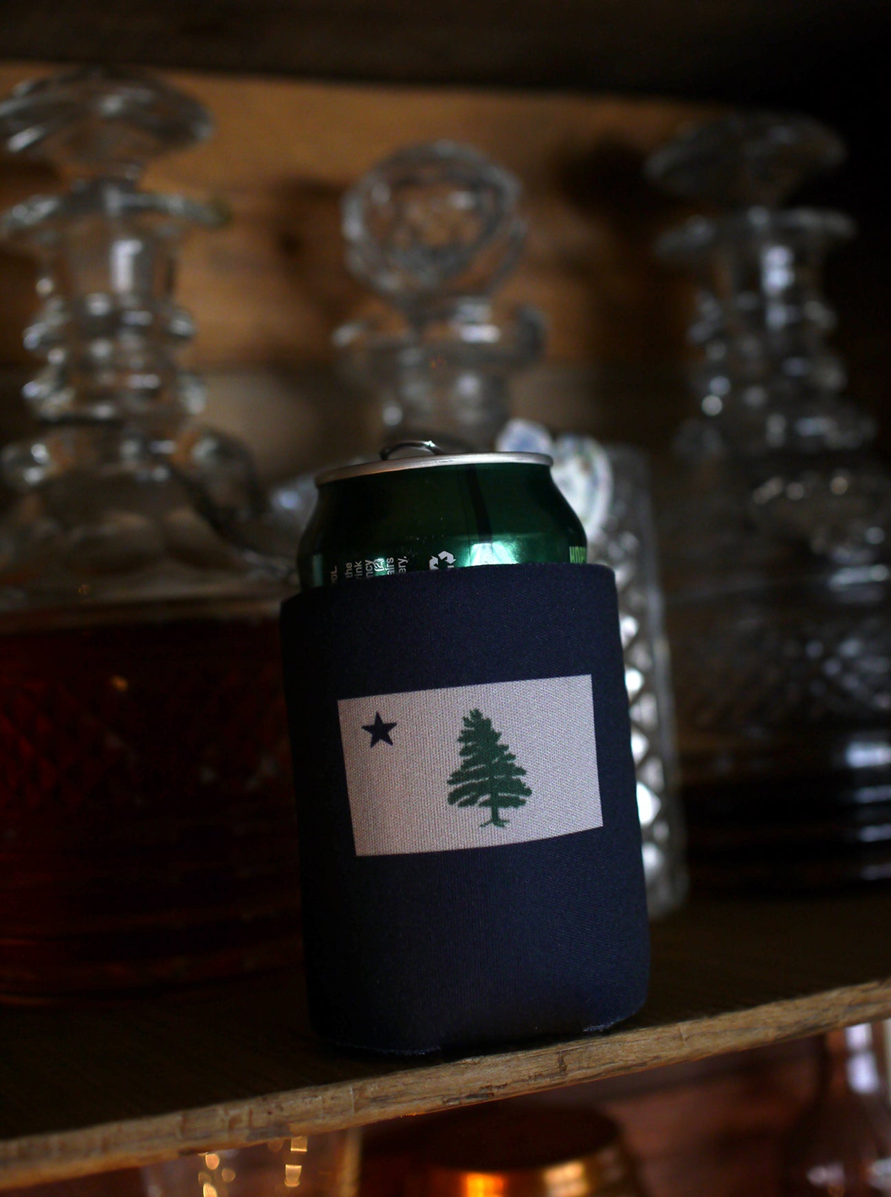 Wicked classy Navy blue can Koozie with two color screen printed image of Original 1901 Maine Flag sitting on a shelf next to antique crystal bottles