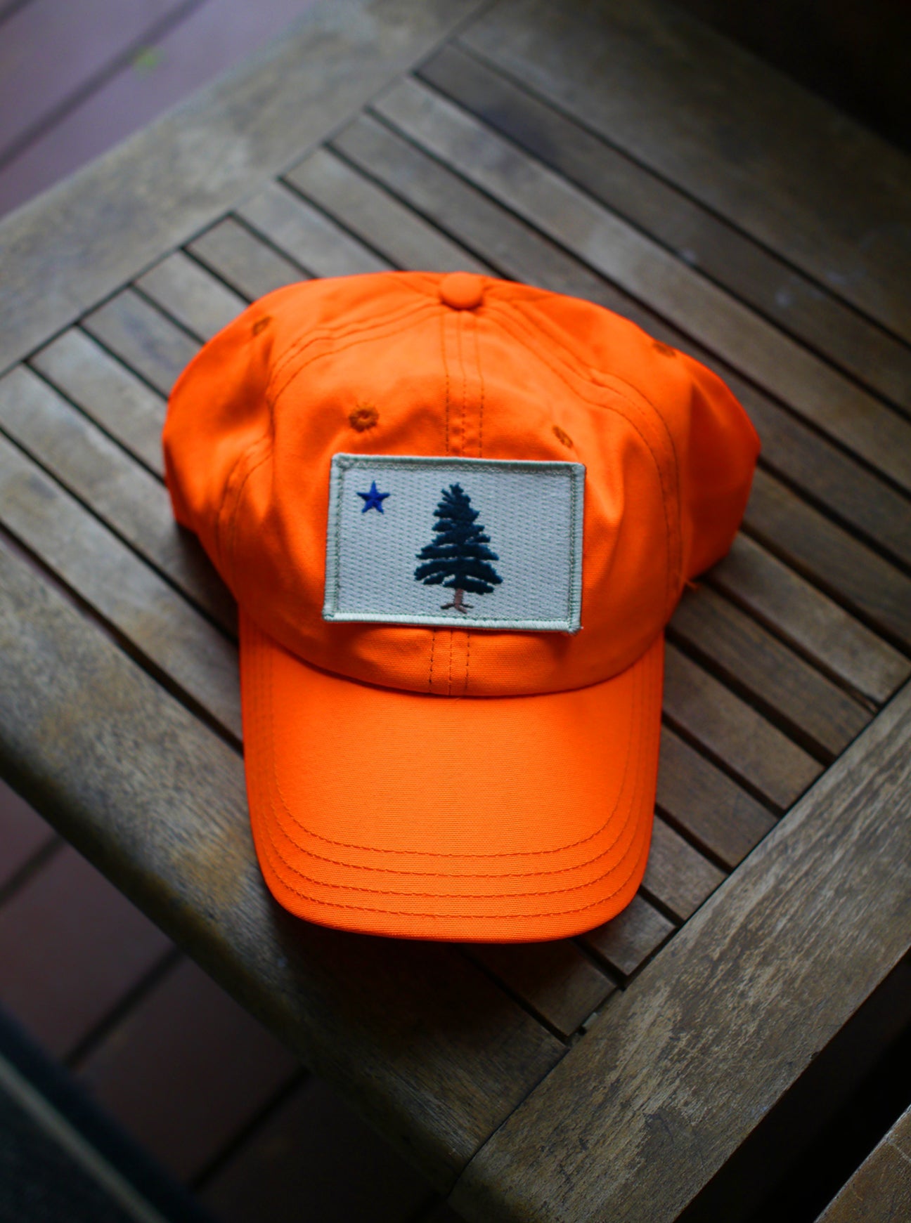 1901 Original Maine Flag insect-repelling baseball cap in hunter safe blaze orange sitting on a wooden bench in the sun. 