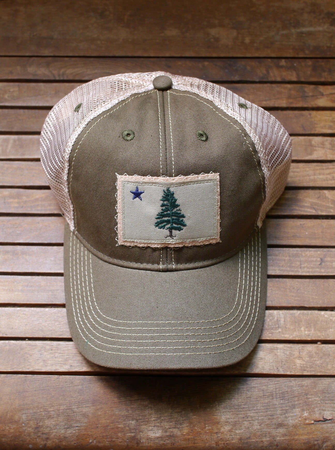 1901 Maine Flag trucker hat is artfully distressed for that lived-in look and feel. Olive trucker hat with tan back mesh and Maine flag appliqué on the front center