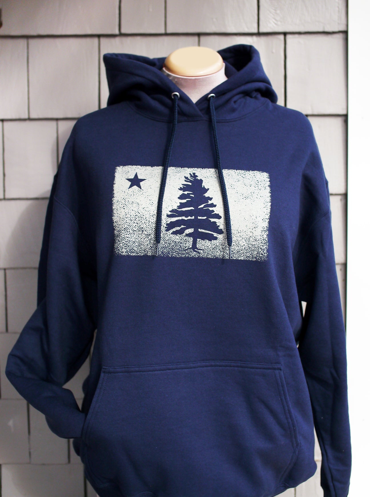 Navy blue hooded sweatshirt features our signature 1901 distressed Maine flag. Sweatshirt is displayed on a mannequin against grey cedar porch shingles..