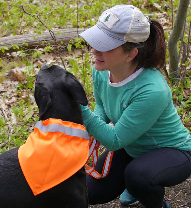 black dog wearing a tick-repelling safety dog collar kerchief. Dog is enjoying an under-chin scratch from his owner, a woman with brown hair wearing a 1901 Maine flag baseball cap